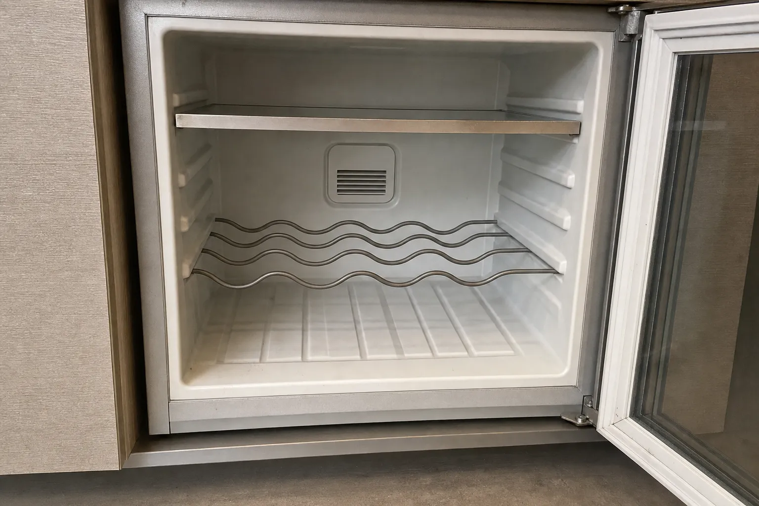 An empty undercounter unit with a wire bottle rack, its glass door swung open